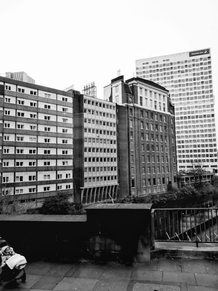 A black and white shot of city centre manchester showing buildings with lots of windows on the banks of the irwell river. A baby being pushed into shot on the left 