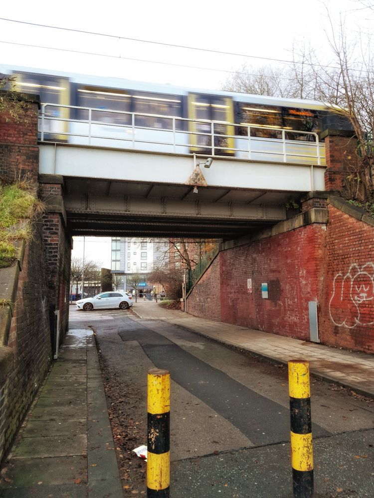 A moving metrolink tram travelling overhead across the Prestwich station bridge into Manchester. Its full of shoppers. In thr foreground are two yellow and black striped bollards