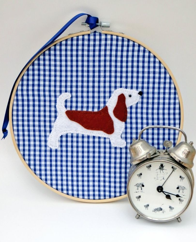 A blue gingham fabric circular embroidery hoop art piece for hanging on the wall by misheleneous. Featuring a felt basset hound dog motif. Displayed alongside a silver metal retro alarm clock