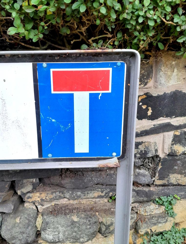 A close up of a red, white and blue Dead End T on a street sign against a stone wall and hedge