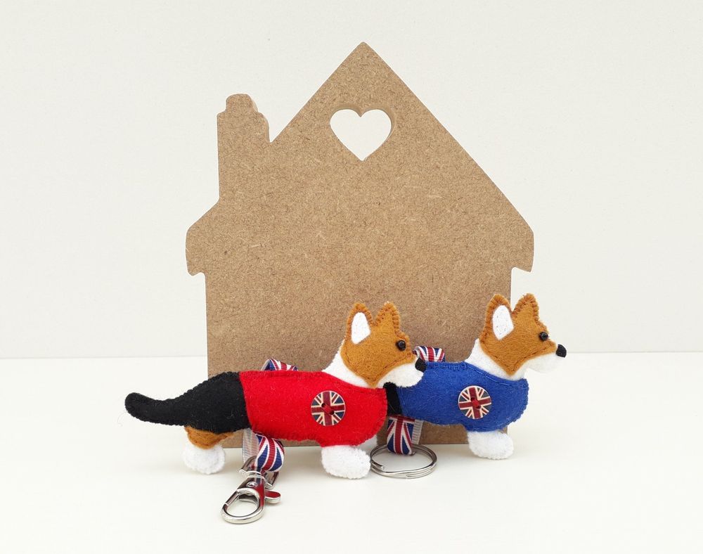 A pair of handmade felt cardigan welsh corgi dog keyrings by misheleneous. Wearing a red and blue jumper with union jack flag motif and matching ribbon. Displayed against a small boat shaped wooden block