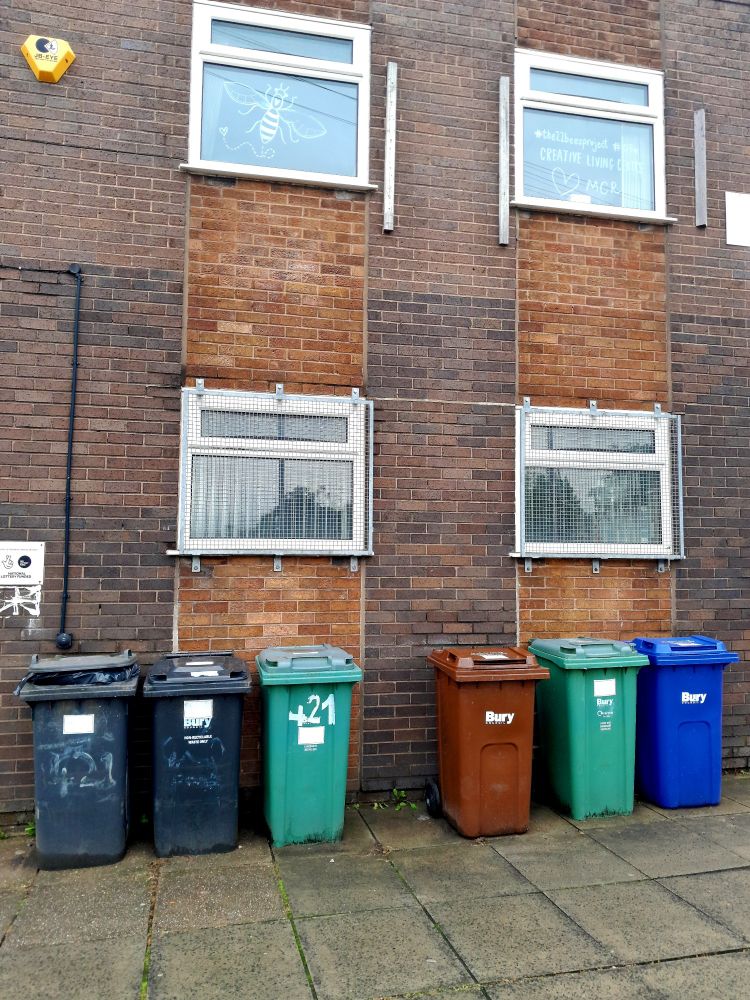 A municipal brown brick building in Prestwich, Manchester, with four square windows. The lower 2 have metal grilles on them. There are 6 wheely bins in various colours arranged against the wall 