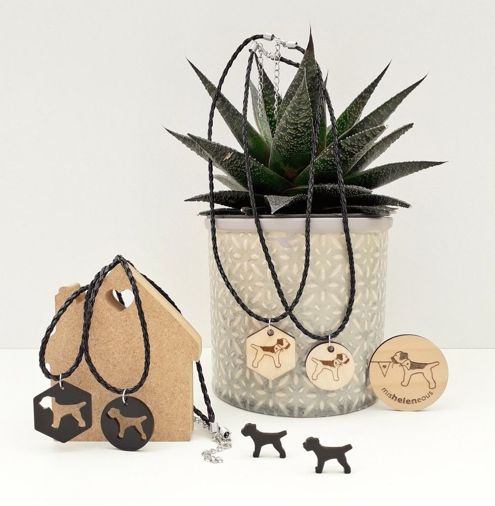 Laser cut black acrylic and engraved finnish ply circular & hexagonal pendants featuring the misheleneous border terrier logo. Hung from faux leather thong with adjustable fastening for unisex wear. Displayed with a potted house plant to the right and a small house shaped wooden block on the left