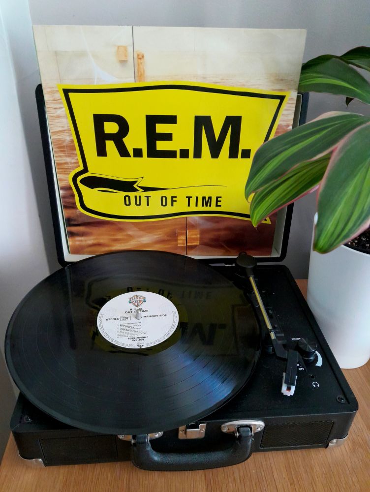 Colour photograph of an orginal copy of vinyl album 'Out of Time' by R.E.M bought in 1991. The cover is resting against the lid of the record player which has the 'memory side' on the turntable which contains my favourite track 'Half a World Away'. There is a houseplant in a pot to the right hand side