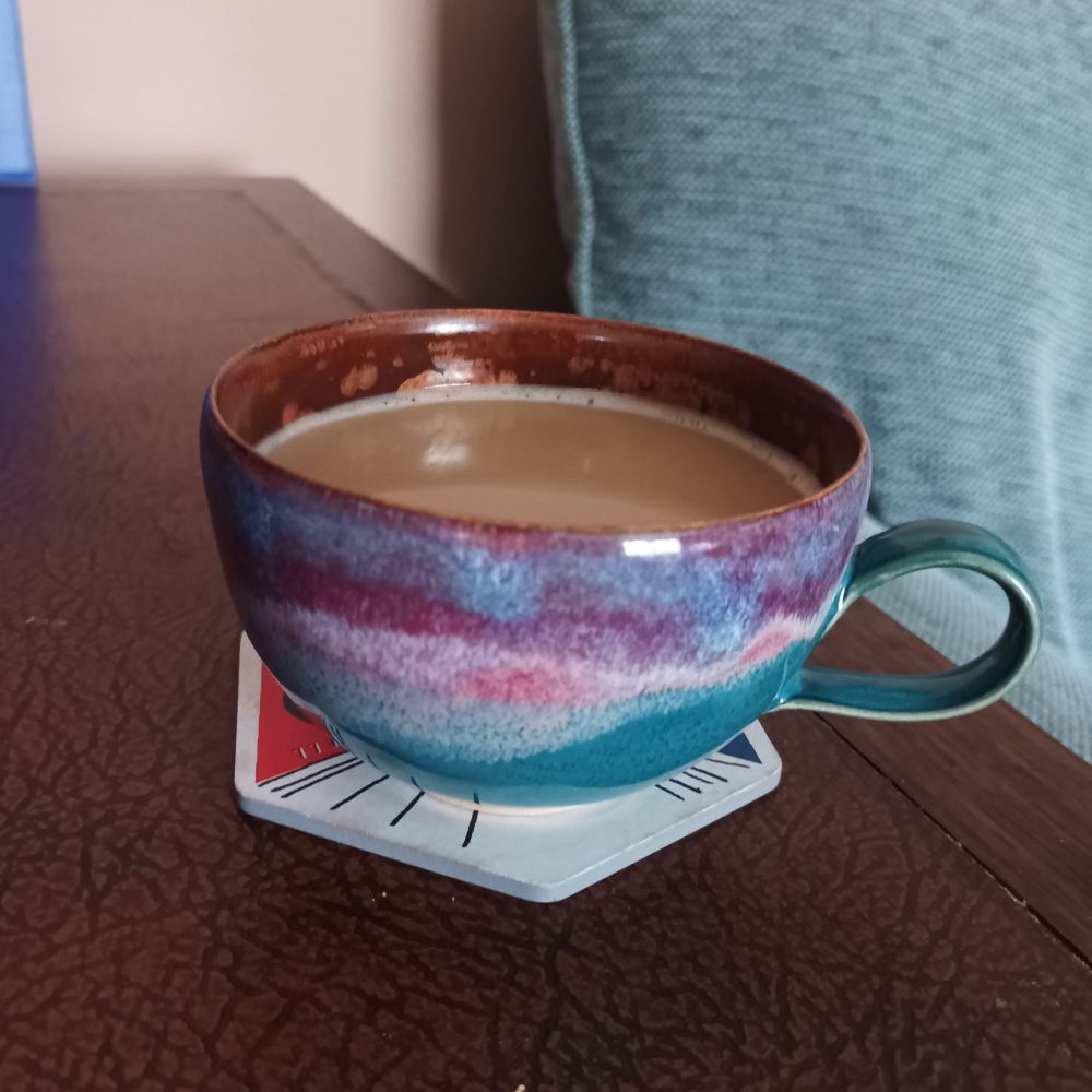 A cup of coffee in a beautiful handmade coffee cup sits on a coaster on a shelf. The cup is particularly pretty with a pink, purple and teal drip glaze on the outside and copper inside. 