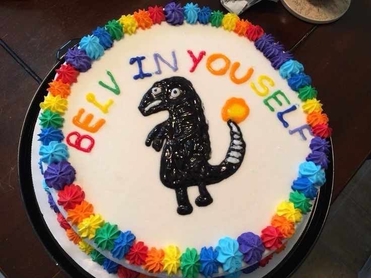 Photo of a poorly decorated cake with rainbow icing surrounding a crude representation of a dinosaur? Kaiju? Anyway, the writing above this weird black blob reads “BELV IN YOUSELF” (sic)