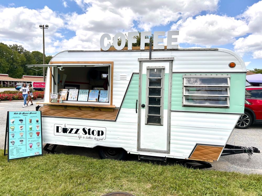 Photograph of a caravan which has been converted into a coffee shop. There is a large serving hatch on the left of frame. There is a pastel blue/green stripe on the right side and two brown, almost quilted, parallelogram shaped patches, one long under the serving hatch, one shorter at bottom right. There is a logo under the hatch that reads “BUZZ STOP - Life is better buzzed” and there’s a sign reading COFFEE  on the roof. An A-board menu stands on the ground on the left of frame. 