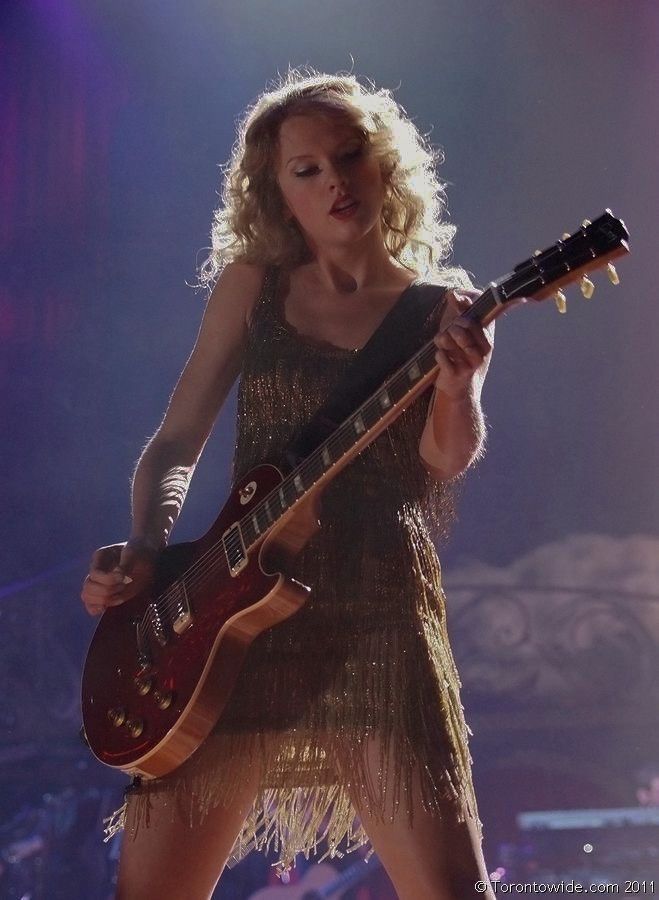 Taylor Swift performing at the Speak Now tour. She is wearing a gold dress and playing a red electric guitar