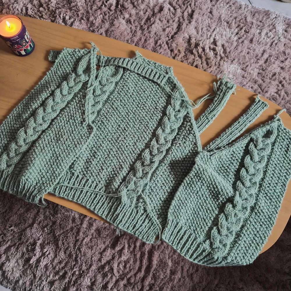 Pieces of a sage green cabled cardigan laying on a coffee table and waiting to be sewed together