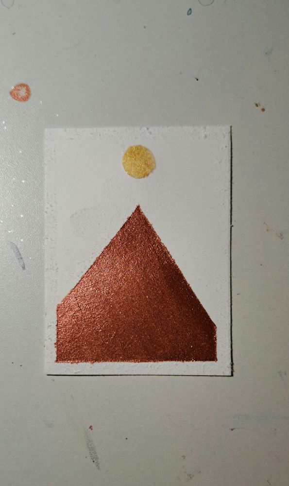A white rectangular piece of white watercolor paper with a metallic red pointed Mountain and a metallic gold Moon directly above the Summit of the mountain