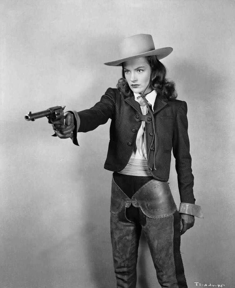 Publicity image of Ella Raines from Tall in the Saddle, against a plain background, dressed in masculine-leaning clothes and a cowboy hat, and holding out a pistol.