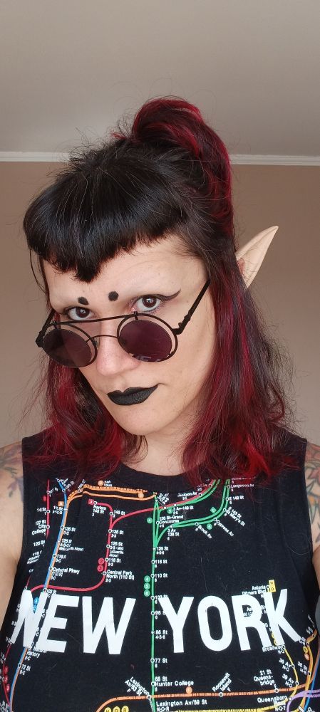 A girl with black and red hair in a bun with bangs, wearing goth makeup, elven ears and a sleeveless shirt with the map of New York Metro.