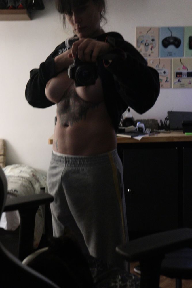 A woman in a dark room lifts her hoodie to show her abs and breasts