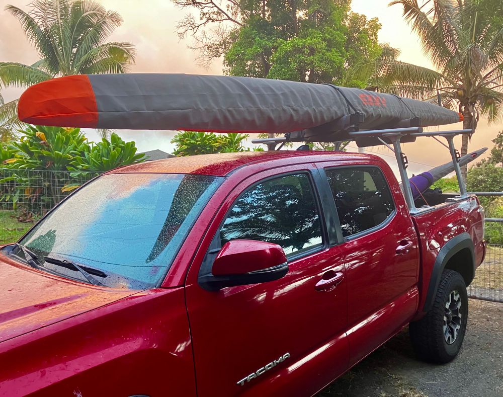 Truck with outrigger canoe on roof rack in a background of ti plants and coconut trees