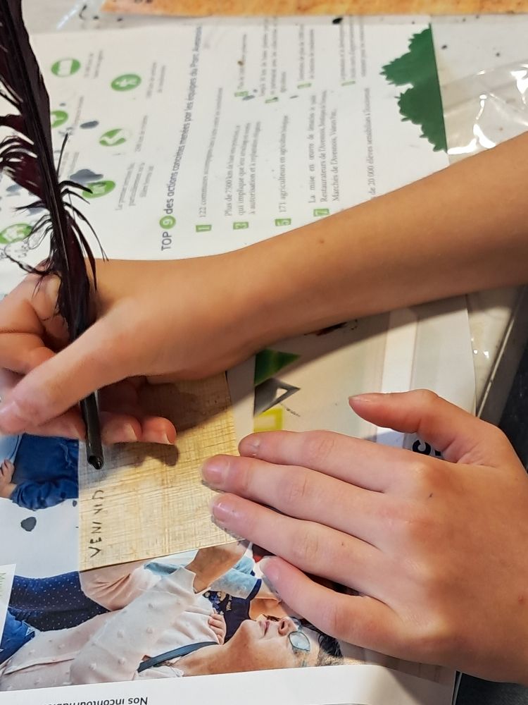 De jeunes mains sont en train d'écrire à la plume sur un petit morceau de papyrus posé sur une feuille de magazine en protection : "Veni vidi...".

Atelier "Écriture en Gaule romaine" au Forum antique de Bavay. 