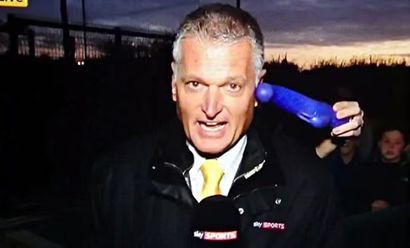 Sky Sports reporter live on Transfer Deadline Day reporting transfer speculation to camera while someone in the crowd places a purple sex toy beside his left ear.