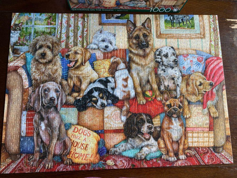 A 1000 piece finished jigsaw puzzle of lots of dogs sitting on and around a sofa. There’s a cushion with “dogs make a house a home” written on it. 