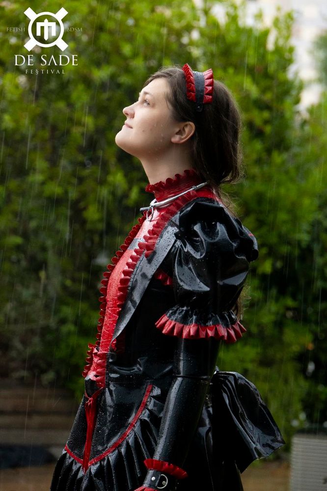 Marii, dressed in her iconic red n black maid dress, stands outside in the rain, looking up