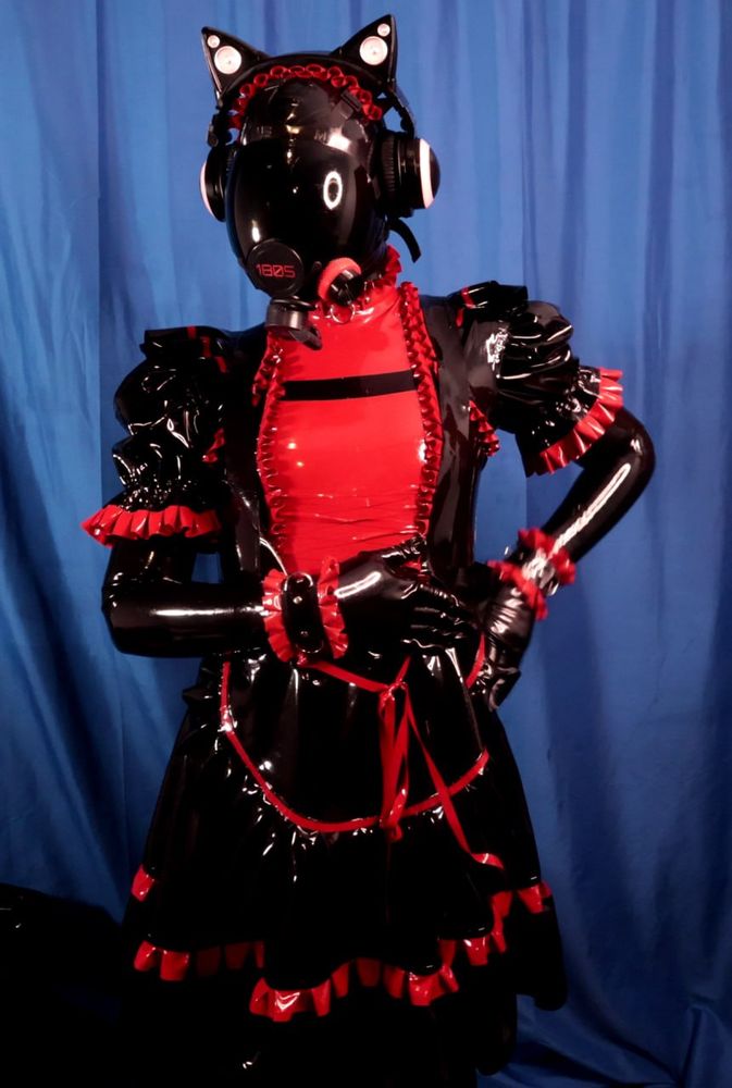 A latex HexDrone with red color theming. It's wearing and red and black maid dress, wrist cuffs, a headband and of course the gasmask. Otherwise it is covered in black rubber. 
Its left hand is on its hip, its right hand is folded across its waist