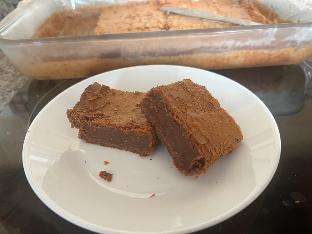 Two brownies arranged on a plate in front of a glass tray containing Yet More brownies