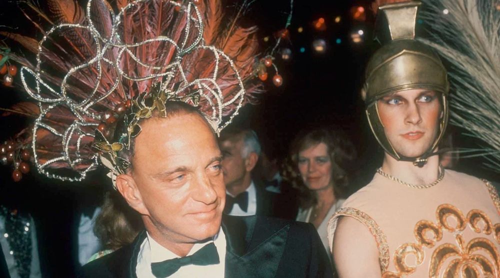 Photo of Roy Cohn in a fabulous sparkly wire & feather headpiece, wearing a tuxedo. Next to him is a young Donald Trump in a bronze-look centurian-style helmet, eye make up & blush, & a singlet w/ a sparkly design. 