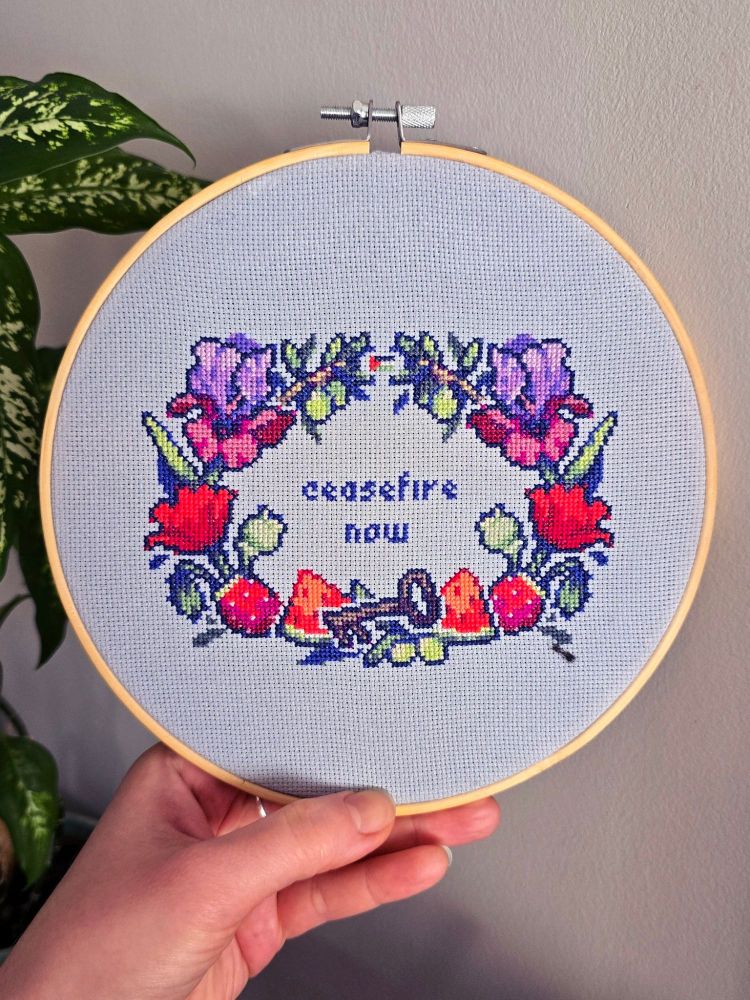 A photo of a cross-stitch being held up in a hoop. The words 'ceasefire now' are surrounded by a border of flowers and objects. The symbolism of the objects chosen, as described by the artist, Ansitru: a key, as symbol of the Nakba; watermelon slices and poppy flowers, which mimic the colors of the Palestinian flag; the Faqqua Iris, Palestine's national flower; and olive branches and a cactus pear, symbols of the Palestinian nation. 