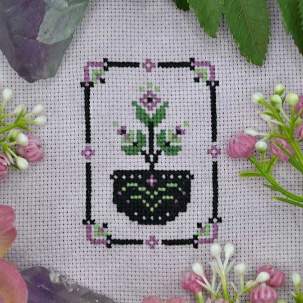 Photo of a flowering cross stitch plant in a geometric style, done on pale purple fabric. The plant is green with purple blooms, and grows in a black pot decorated with green stripes and purple flower motifs. Around the plant is a matching border. The cross-stitch is displayed with real-life buds and leaves around it. 