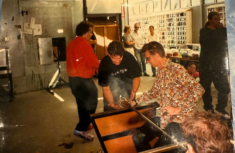 David Byrne sits at a glass blowing bench in a brightly colored shirt while Ashley Rowley, in a black Go Outside shirt, leans over to help him shape the hot glass bubble. In the background in the glass studio, a handful of onlookers sit or stand in front of shelving full of containers of brightly colored glass frit while Dale Chihuly walks by in a bright red shirt. 
