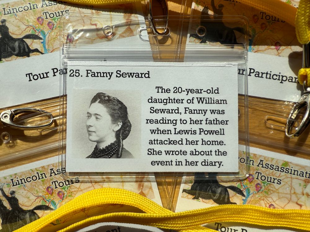 A photograph of a tour badge and lanyard laying on top of other tour badges. The image in the main badge is a photograph of Fanny Seward. Her name is at the top followed by the words “The 20-year-old daughter of William Seward, Fanny was reading to her father when Lewis Powell attacked her home. She wrote about the event in her diary.” 