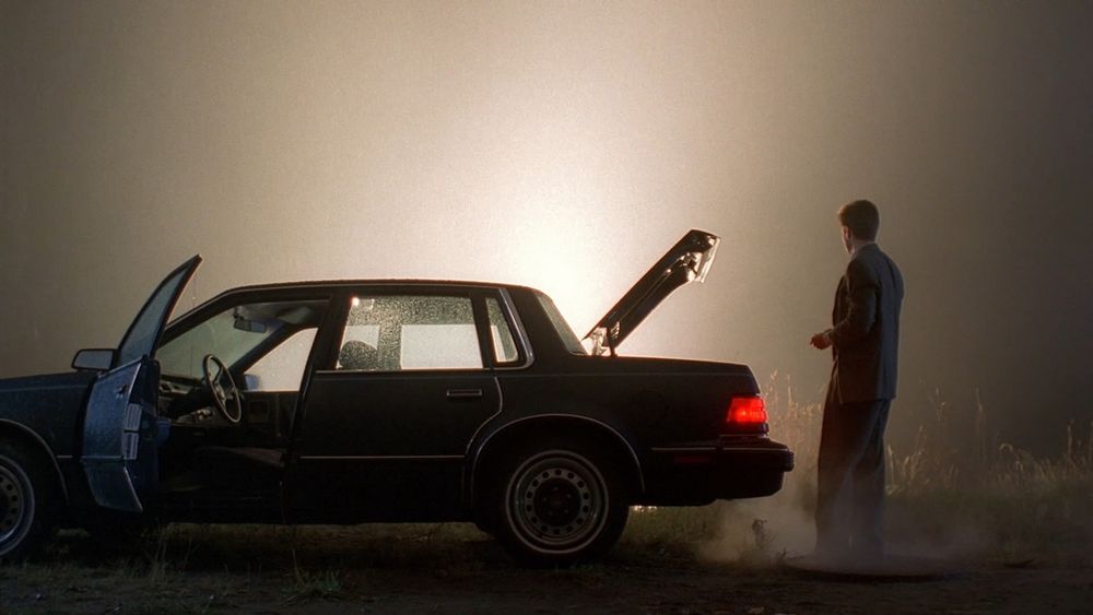 Mulder stands at a car, with an open door and open boot, as a bright light rises behind them. 