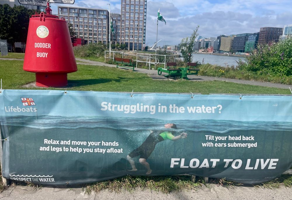 photo of a large roadside banner beside the dodder buoy in Dublin. by RNLI lifeboats, it shows a person wearing a wetsuit, floating with their head above the water. prompt is, struggling in the water? and the instructions are: float to live; relax and move your hands and legs to help you stay afloat; tilt your head back with ears submerged