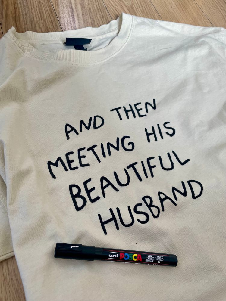Photo of a white t-shirt with black hand lettering that reads “and then meeting his beautiful husband”, with a black posca marker sitting on the shirt. 