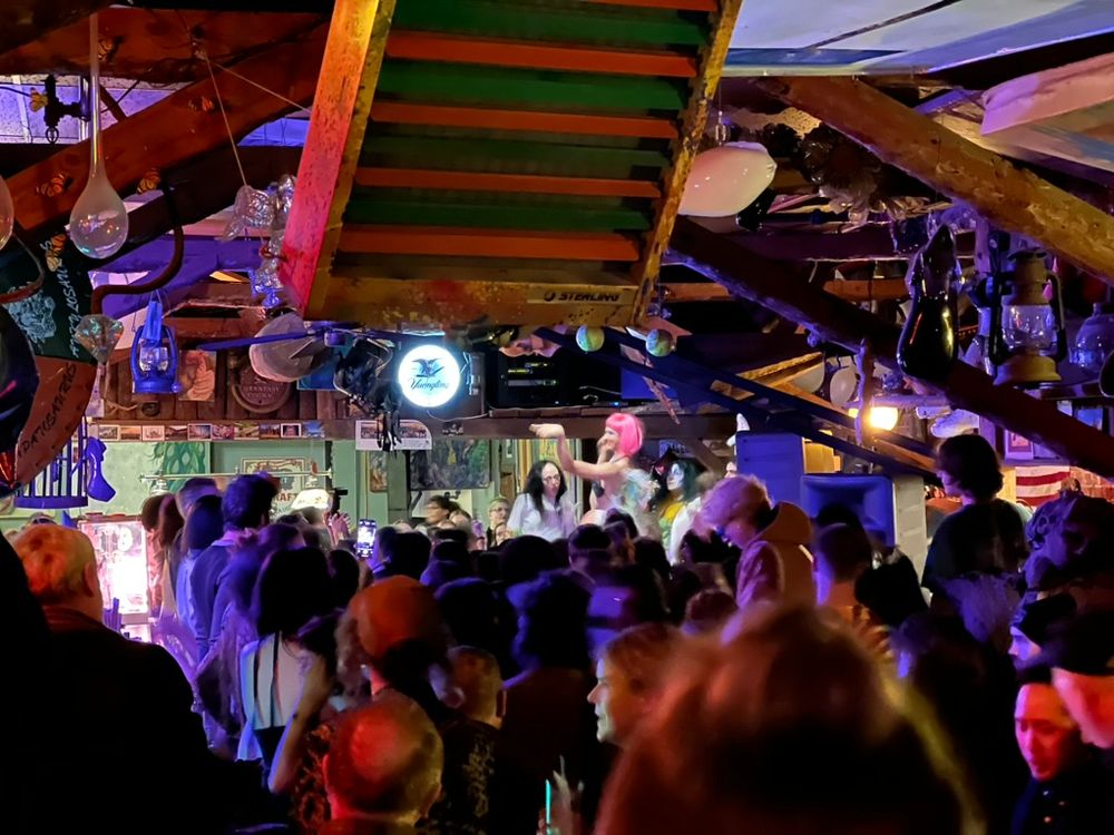 A shot over the head of the crowd in a small bar covered in art and with lanterns and other glasswork hanging from the ceiling. In the background, drag performers dance in the background. A slight motion blur on the image and the colored lights reflecting off the many surfaces create a dreamy feeling.