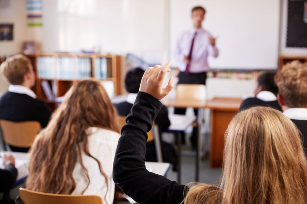 A classroom with pupils at desks listening to a male teacher. A female pupil with long hair has her hand raised to ask a question.