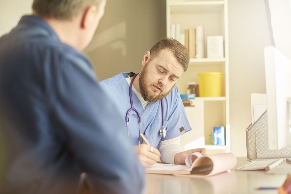 A male doctor is writing notes in a consultation with a male patient.