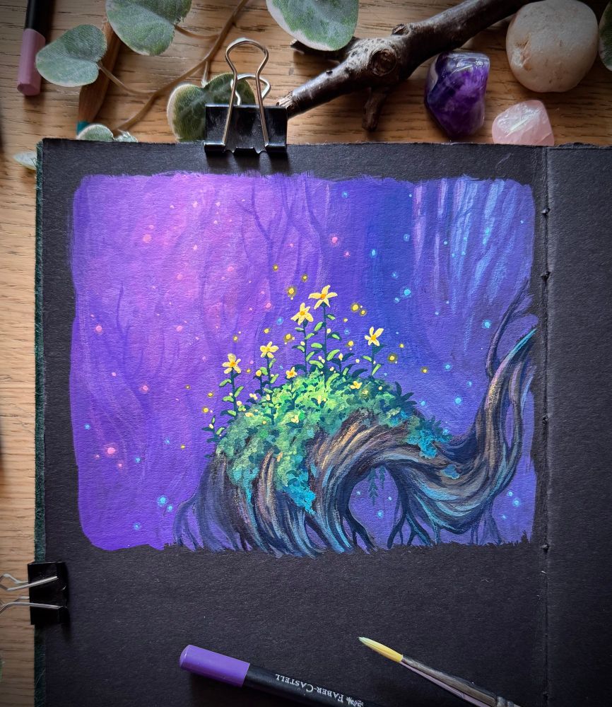 A photo of the left page of an open, black paper sketchbook, placed on a wooden desk and surrounded by coloured pencils, a paint brush, leaves, a branch, and minerals. The sketchbook page features part of a smoothly curving tree, overgrown with moss and bright yellow flowers. In the background, a pink/purple/blue forest is visible indistinctly. Pollen and shimmering dust dance in the air.