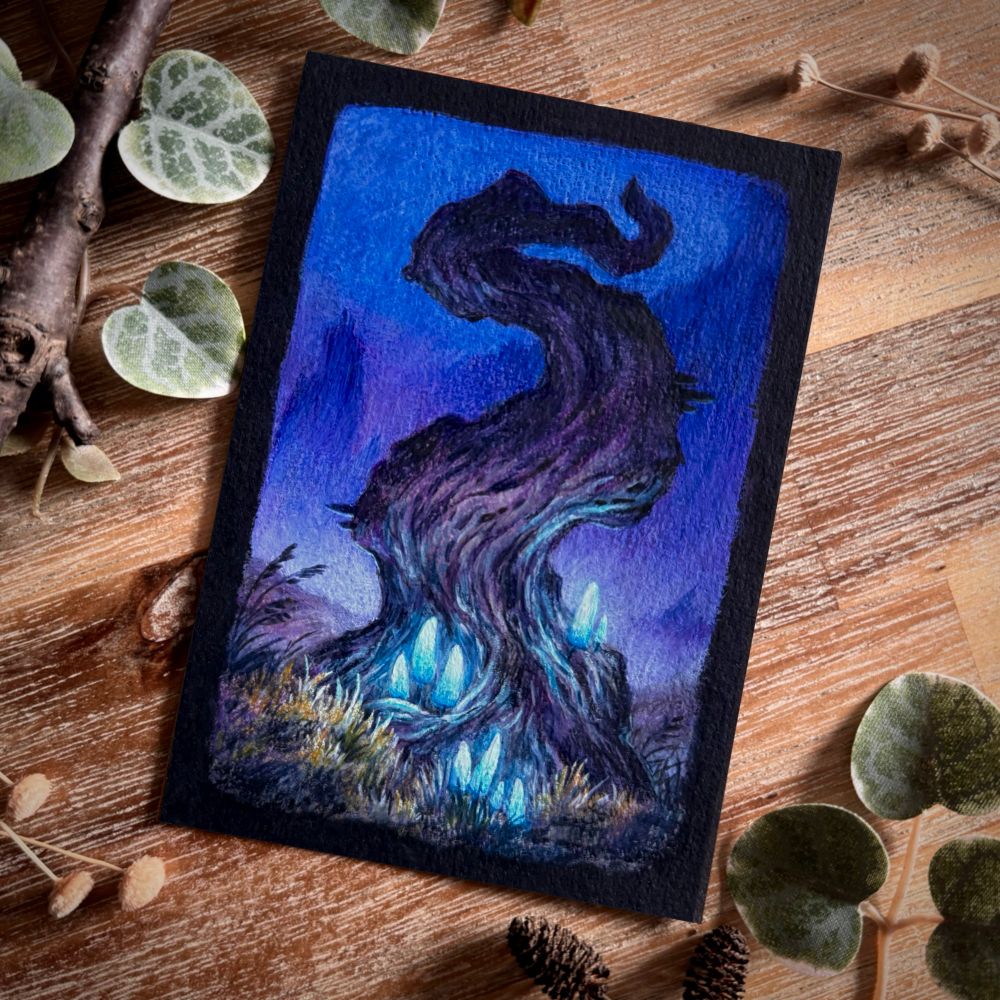 A photo of a painting on black paper, placed on a wooden table and surrounded by branches, dried flowers, and leaves for decoration. The painting features a twisting, bare tree with pale blue glowing mushrooms at its base. Dry yellowish grass surrounds it and indistinct mountain-like shapes are visible on the dark purple background.