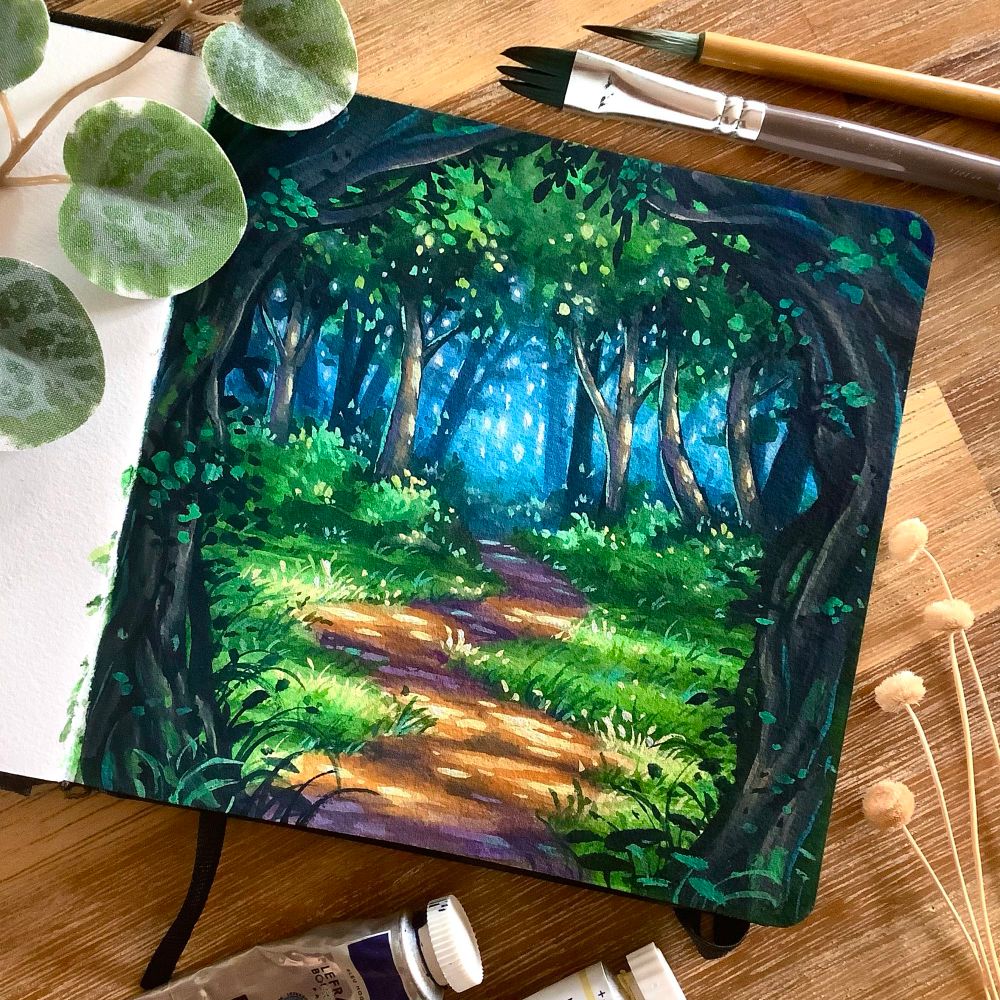 A photo of an open square sketchbook placed on a wooden table, surrounded by art supplies and plants for decoration. The sketchbook page features pathway with dappled light leading over a small clearing in the forest, a gnarled wooden portal framing the front, and bright blue woods visible in the back.