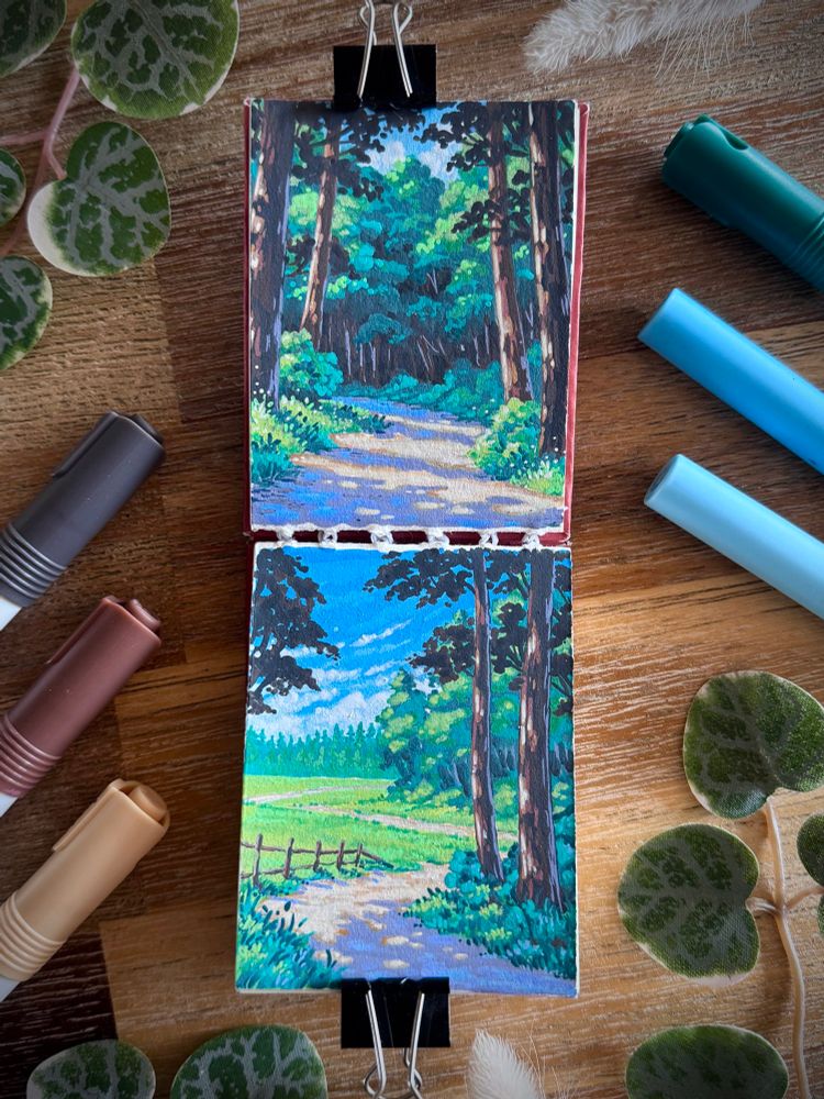 A photo of an open sketchbook placed on a wooden table and surrounded by acrylic markers and leaves for decoration. Both sketchbook pages feature summery landscapes with dappled light, the top one of a pathway in the forest, the bottom one of a pathway leading out of a forest towards grassy fields.