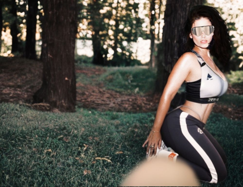 I'm wearing Oakley sunglasses and activewear posing on grass in a outdoor setting which has a lot of woods nearby during evening/dusk