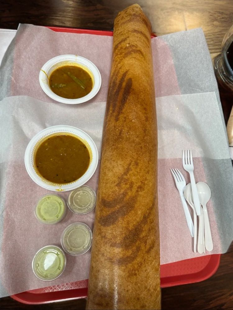 An extra large masala dosa 2 mint chutney, 2 groundnut and coconut chutney kept in a lid tight cup, 2 plates of sambar and 2 disposable spoon and forks kept on food paper served on a red plate