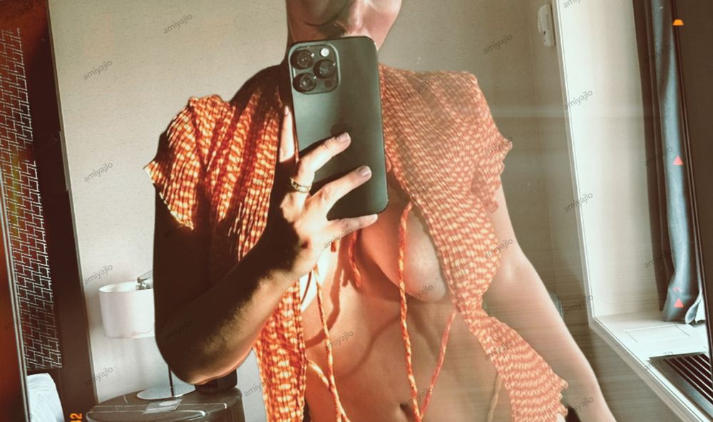 A person takes a mirror selfie in a windowside lit room, wearing an orange top and beige bottoms