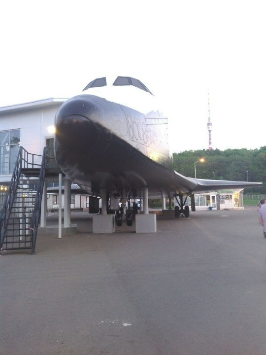 A space shuttle orbiter displayed outdoors on concrete supports near a building, with stairs and greenery in the background.