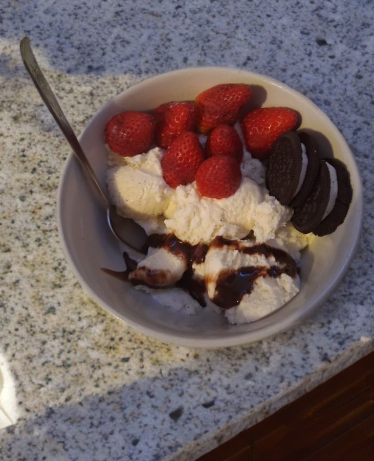A bowl of vanilla ice cream topped with fresh strawberries, chocolate sandwich cookies, and chocolate syrup, with a spoon on the side.