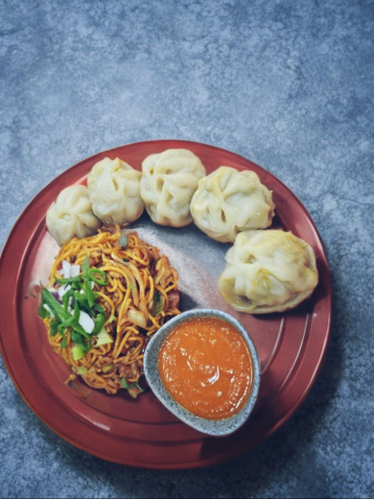 5pcs of momos, with chicken chowmein with a bowl of chutney served on a red plate.