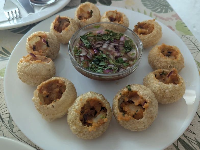 A plate of filled golgappa (pani puri) arranged in a circle around a bowl of spicy water with chopped onions and herbs.