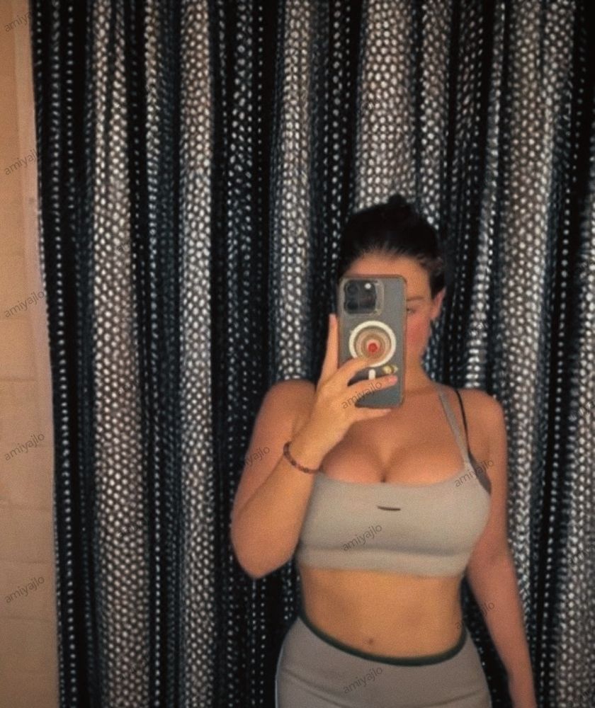 A person in a gray sportswear and shorts takes a mirror selfie, holding a phone in front of their face. Patterned curtains are visible in the background.