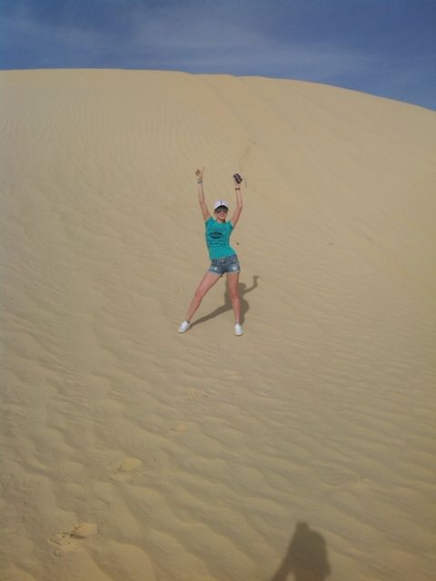 Standing with my arms raised on a sandy dune under a blue sky, wearing a teal shirt, shorts, white shoes, and a white cap.