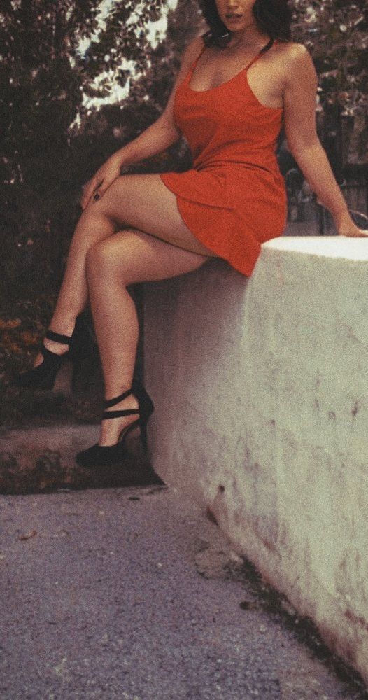 Wearing a red dress and black shoes sits on a low white wall outdoors, with trees and plants in the background.