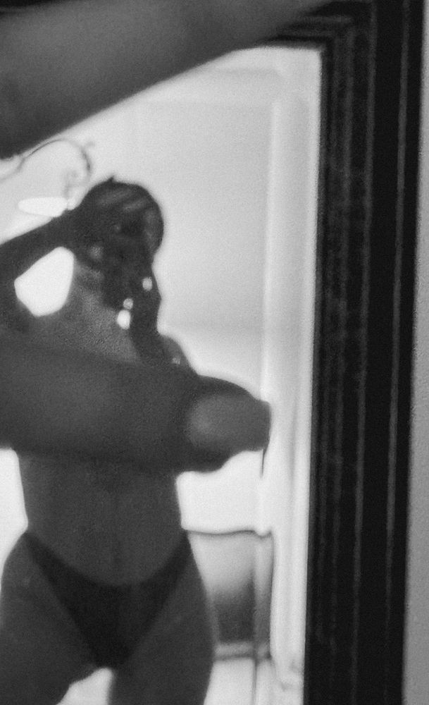 Black and white photo of a person in underwear taking a mirror selfie; the image is blurred and partially obscured by an arm in the foreground.